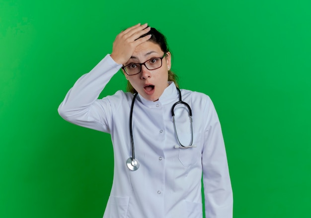 Verrast jonge vrouwelijke arts die medische mantel en stethoscoop en bril draagt die hand op hoofd houdt dat op groene muur met exemplaarruimte wordt geïsoleerd