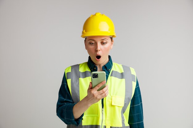 Verrast jonge vrouwelijke bouwvakker die veiligheidshelm en veiligheidsvest draagt ?? die en mobiele telefoon bekijkt