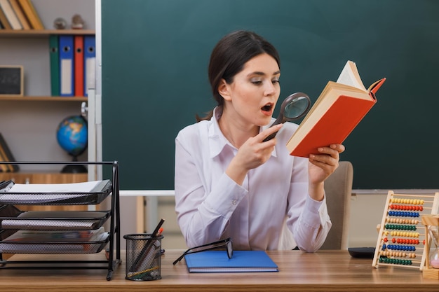 Verrast jonge vrouwelijke leraar leesboek met vergrootglas zit aan bureau met schoolhulpmiddelen in de klas