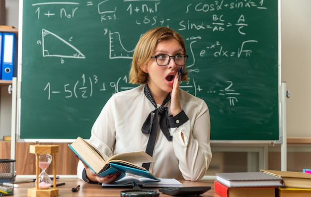 Verrast jonge vrouwelijke leraar met een bril zit aan tafel met schoolhulpmiddelen die een boek vasthouden en de hand op de wang in de klas leggen