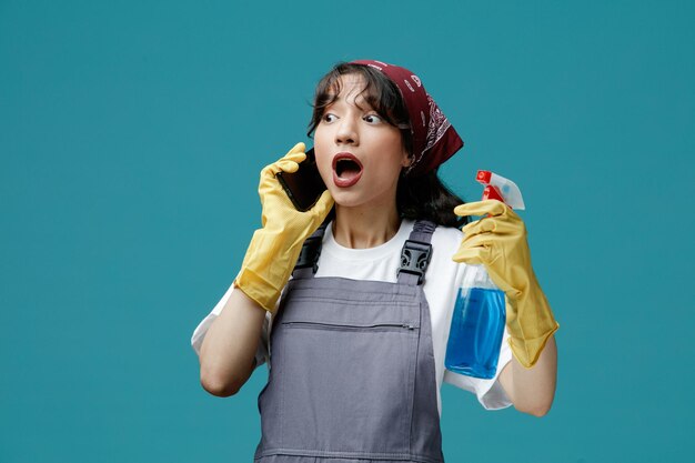 Verrast jonge vrouwelijke schoonmaker met uniforme bandana en rubberen handschoenen met reinigingsmiddel kijkend naar kant pratend over telefoon geïsoleerd op blauwe achtergrond