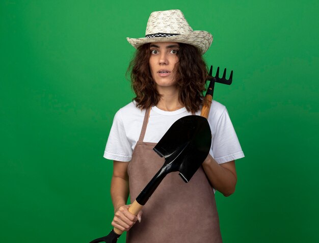 Verrast jonge vrouwelijke tuinman in uniform dragen tuinieren hoed bedrijf spade en hark op schouder zetten
