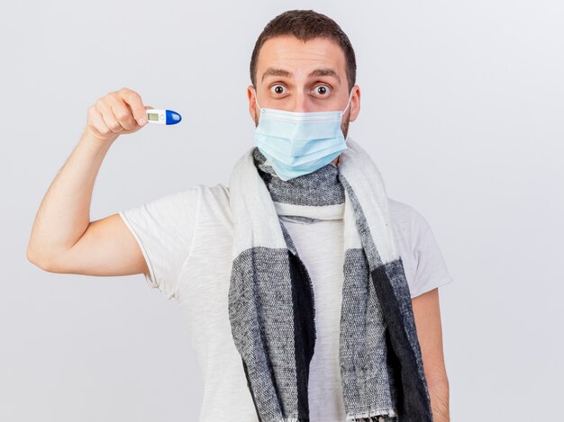 Verrast jonge zieke man met medische masker en sjaal houden thermometer geïsoleerd op een witte achtergrond