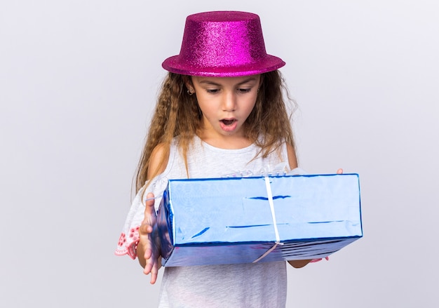 Verrast klein kaukasisch meisje met paarse feestmuts die een geschenkdoos vasthoudt en kijkt op een witte muur met kopieerruimte