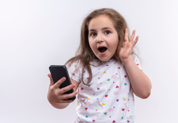 Verrast klein schoolmeisje die de witte telefoon van de t-shirtholding dragen die luistergebaar op geïsoleerde witte achtergrond toont