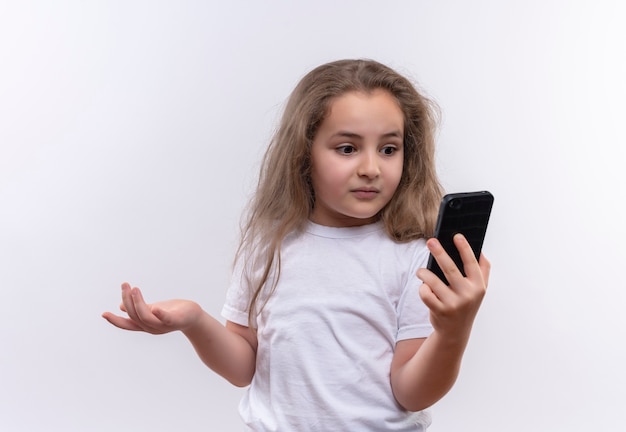 Verrast klein schoolmeisje die witte t-shirt dragen die op telefoon op geïsoleerde witte achtergrond kijken