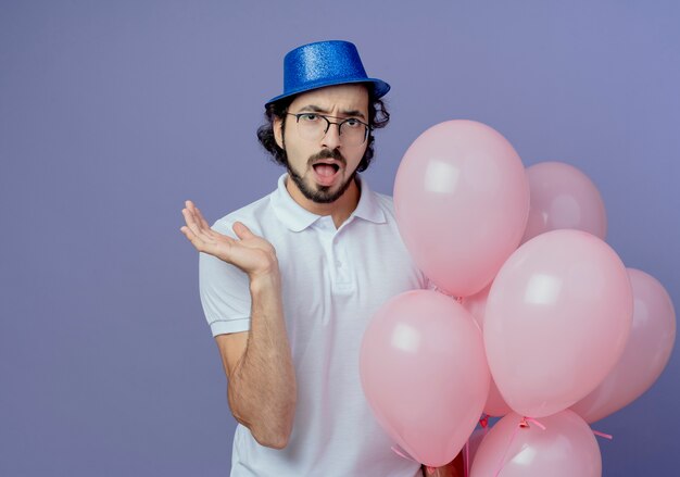 Verrast knappe man met bril en blauwe hoed met ballonnen en verspreid hand geïsoleerd op paarse achtergrond