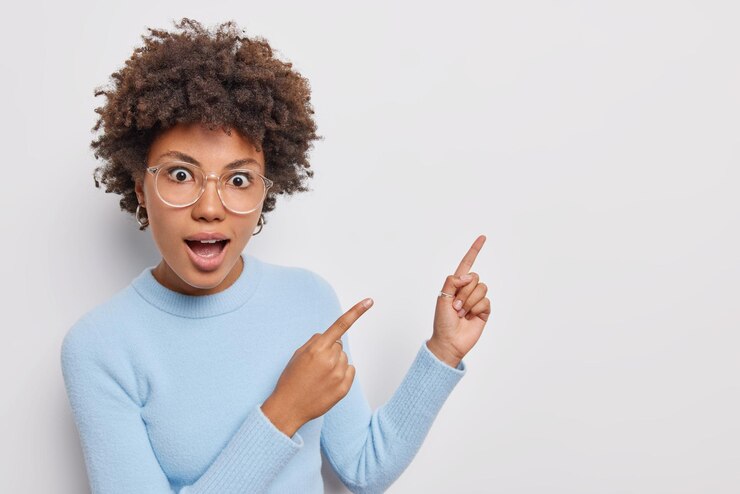 Gratis Foto verrast krullend haired jonge vrouw wijst in de rechterbovenhoek toont mooie advertentie onverwachte aanbieding of korting draagt grote transparante bril blauwe coltrui geïsoleerd op witte achtergrond