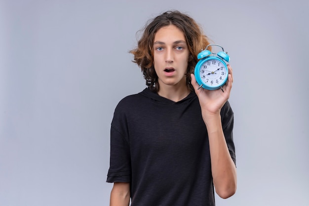 Verrast man met lang haar in zwart t-shirt met een wekker op witte achtergrond