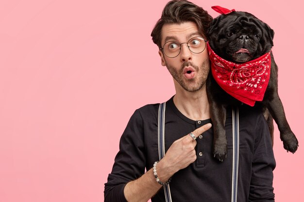 Verrast mannelijke hipster gekleed in een modieus zwart shirt, wijst naar zijn grappige hond met rode bandana, voelt zich verrast zoals gekocht voor een lage prijs, geïsoleerd over roze muur met lege ruimte aan de linkerkant