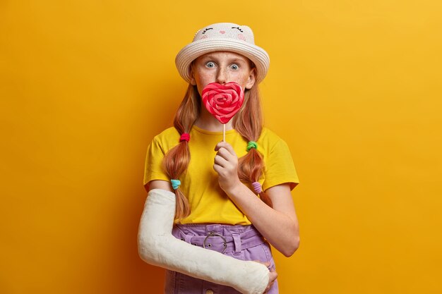 Verrast meisje bedekt mond met grote lolly, geniet van zomertijd, zoetekauw en houdt heerlijk snoep, gekleed in stijlvolle outfit, heeft gebroken arm, geïsoleerd op gele muur.