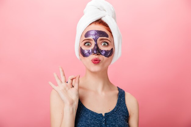 Verrast meisje dat goed teken toont tijdens huidverzorgingsbehandeling. Vooraanzicht van verbaasde vrouw met gezichtsmasker geïsoleerd op roze achtergrond.