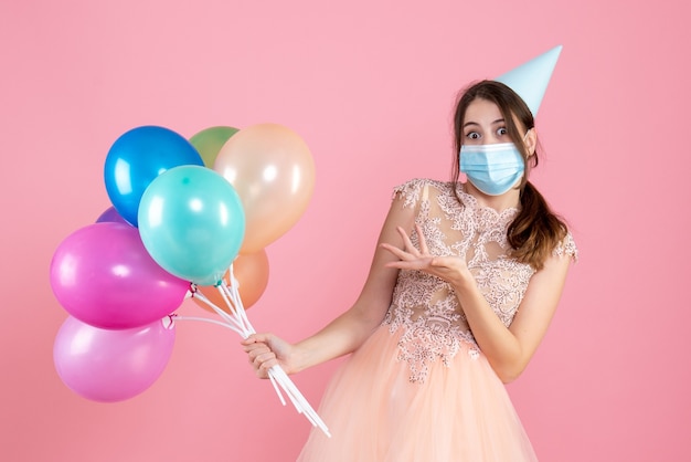 verrast meisje met feestmuts en medisch masker met kleurrijke ballonnen op roze