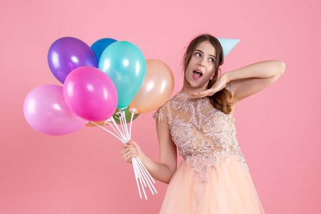 verrast meisje met feestmuts met ballonnen op roze