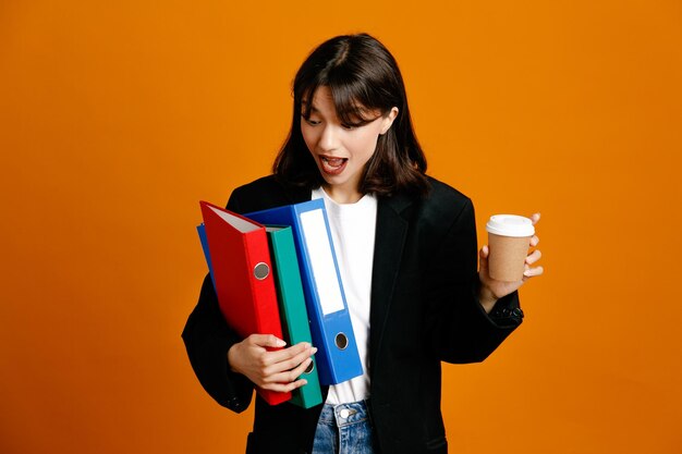 Verrast met mappen met koffiekopje jonge mooie vrouw draagt zwarte jas geïsoleerd op oranje achtergrond