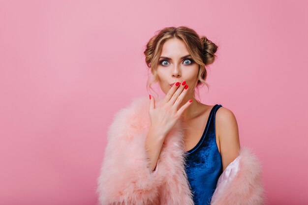 Verrast mooi meisje met schattig kapsel zei iets verkeerds, terwijl ze poseerde voor een roze muur. Schattige jonge vrouw in fluwelen bodysuit bedek haar mond met hand, geïsoleerd op lichte achtergrond