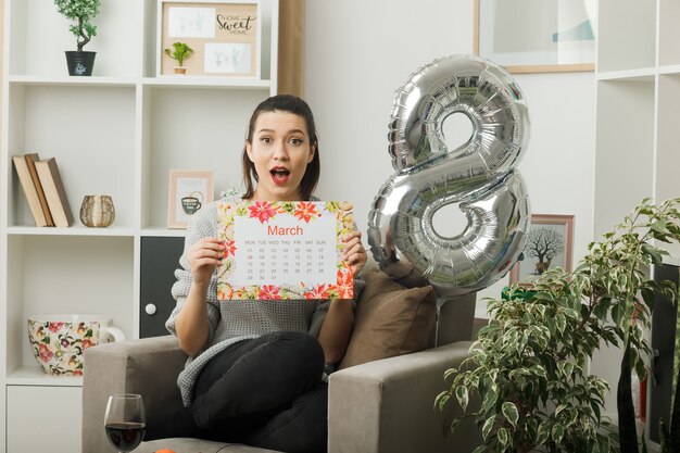 Verrast mooi meisje op gelukkige vrouwendag met kalender zittend op een fauteuil in de woonkamer