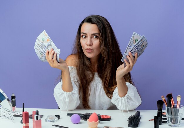 Verrast mooi meisje zit aan tafel met make-up tools geld in twee handen houden geïsoleerd op paarse muur