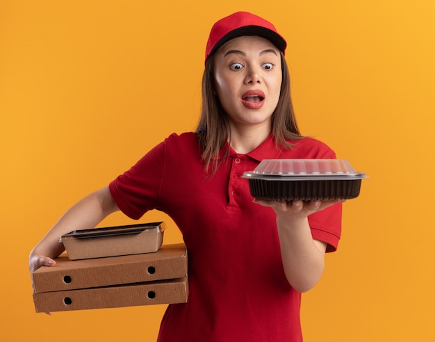Verrast mooie bezorger in uniform houdt papieren voedselpakket op pizzadozen en kijkt naar voedselcontainer geïsoleerd op oranje muur met kopieerruimte