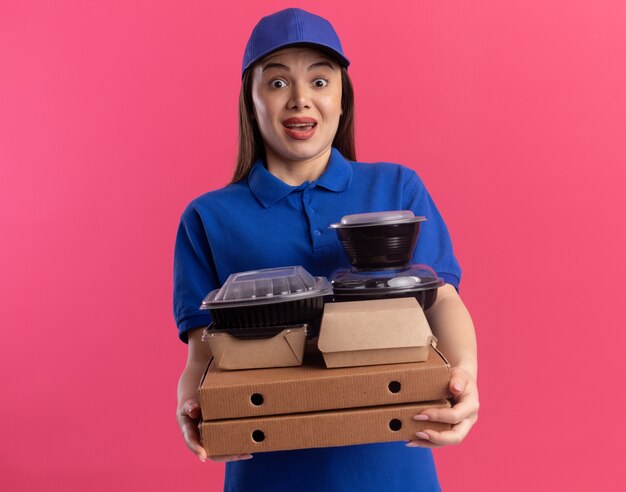 Verrast mooie bezorger in uniform houdt voedselpakket en containers op pizzadozen en kijkt naar de camera op roze