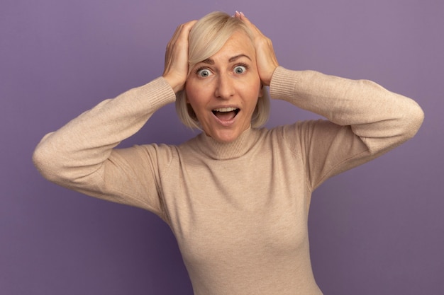 Verrast mooie blonde slavische vrouw legt handen op het hoofd geïsoleerd op paarse muur