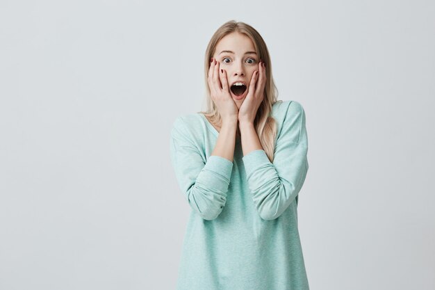 Verrast mooie jonge vrouw met lang geverfd blond haar, kijkt met geopende mond, geschokt om iets vreselijks te zien, terloops gekleed. Mensen, verrassing, gezichtsuitdrukkingen