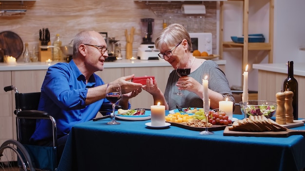 Verrast oma kijken naar cadeau van grootvader geven tijdens feestelijk diner. geïmobiliseerde, verlamde, gehandicapte bejaarde man die thuis met zijn vrouw dineert, van de maaltijd geniet, hun verjaardag viert