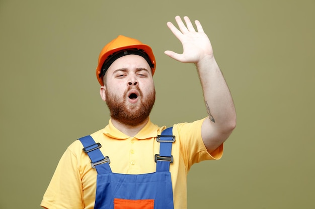 Gratis foto verrast opgeheven hand jonge bouwer man in uniform geïsoleerd op groene achtergrond