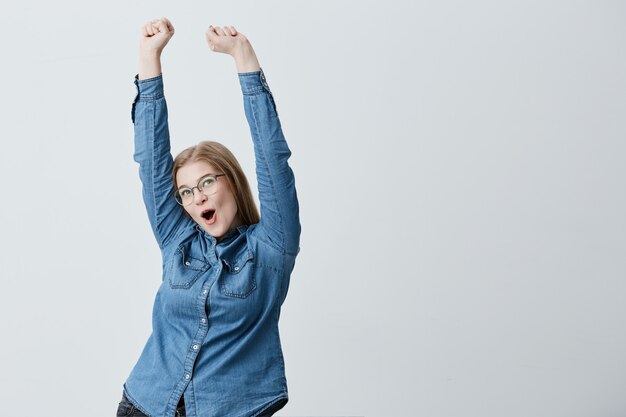 Verrast opgewonden jonge vrouw met blond steil haar, bril, in spijkerblouse heft armen in de lucht en viert haar triomf. Gelukkig blonde jong wijfje dat haar succes verheugt.