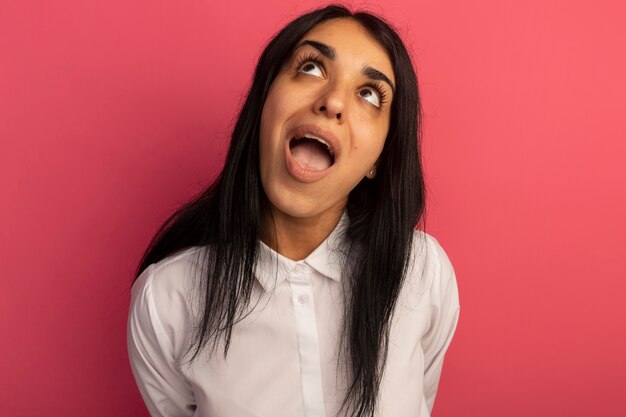 Verrast opzoeken van jong mooi meisje met witte t-shirt geïsoleerd op roze