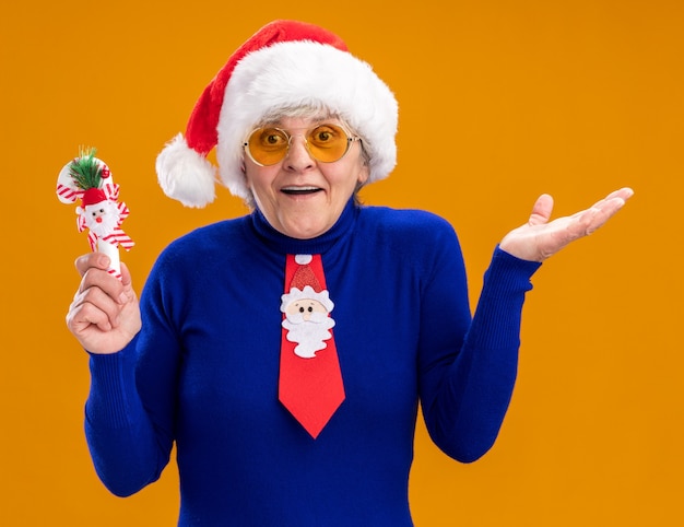 Verrast oudere vrouw in zonnebril met kerstmuts en kerstman stropdas houdt riet van het suikergoed en houdt de hand open geïsoleerd op een oranje achtergrond met kopie ruimte