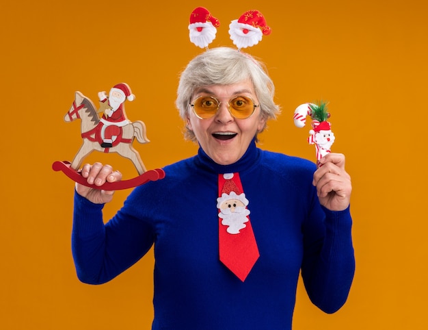 Verrast oudere vrouw in zonnebril met santa hoofdband en santa stropdas santa houden op hobbelpaard decoratie en riet van het suikergoed geïsoleerd op een oranje achtergrond met kopie ruimte