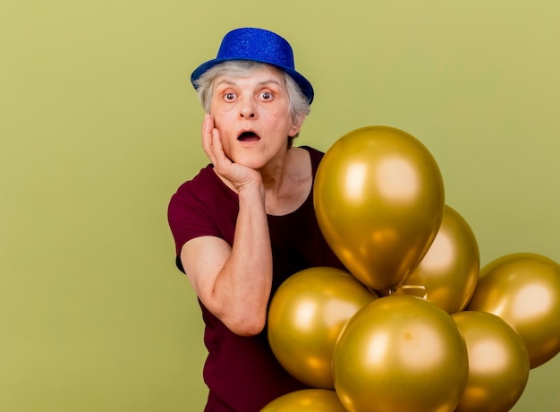 Verrast oudere vrouw met feestmuts legt hand op kin en houdt helium ballonnen geïsoleerd op olijfgroene muur met kopie ruimte