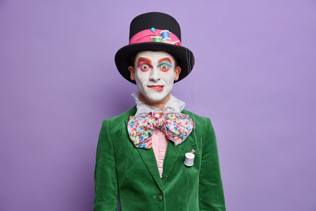 Verrast parade festival deelnemer heeft afbeelding van hoedenmaker uit wonderland draagt lichte make-up gekleed in carnavalskostuum vormt tegen levendige paarse muur