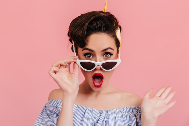 Verrast pinupmeisje met rode lippen wat betreft zonnebril. Studio shot van verbaasde elegante vrouw geïsoleerd op roze achtergrond.