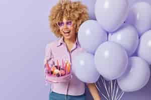 Gratis foto verrast positieve vrouw met krullend haar houdt bos van opgeblazen ballonnen en verjaardagstaart viert verjaardag of speciale gelegenheid geïsoleerd over paarse achtergrond lege ruimte voor reclame