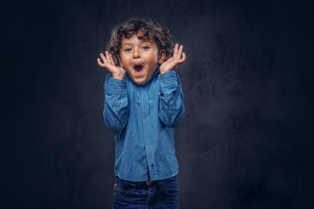 Verrast schattige schooljongen met bruin krullend haar gekleed in een blauw shirt, poseren in een studio. Geïsoleerd op een donkere gestructureerde achtergrond.