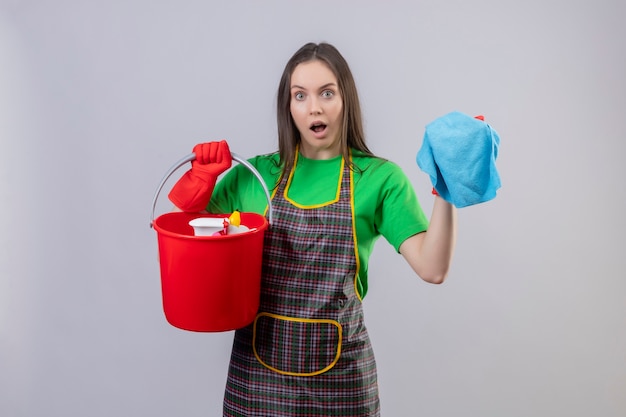 Verrast schoonmakend jong meisje die uniform in rode handschoenen dragen die schoonmakende hulpmiddelen en doek op geïsoleerde witte achtergrond houden