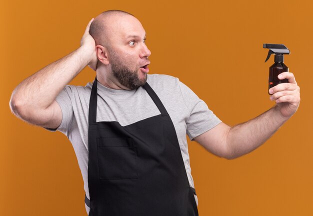 Verrast Slavische mannelijke kapper van middelbare leeftijd in uniform vasthouden en kijken naar waternevelfles hand zetten achter hoofd geïsoleerd op oranje muur