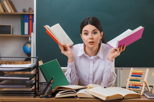 verrast verspreidende handen jonge vrouwelijke leraar met boek zittend aan bureau met schoolhulpmiddelen in de klas