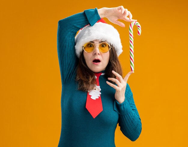 Verrast volwassen blanke vrouw in zonnebril met kerstmuts en stropdas van de kerstman bedrijf candy cane met vingers geïsoleerd op een oranje achtergrond met kopie ruimte