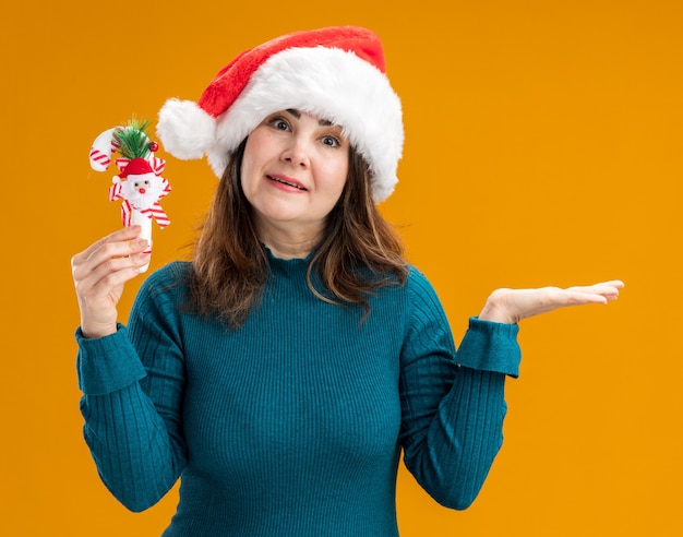 verrast volwassen blanke vrouw met kerstmuts houdt riet van het suikergoed en houdt de hand open geïsoleerd op een oranje achtergrond met kopie ruimte