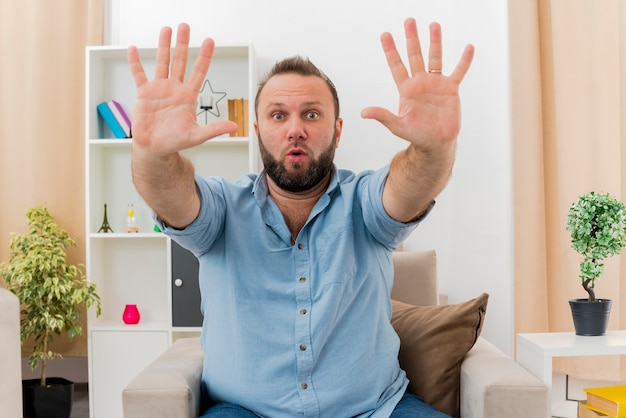 Gratis foto verrast volwassen slavische man zit op een fauteuil met twee handen in de woonkamer