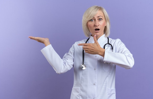 Verrast volwassen vrouwelijke arts in medisch gewaad met stethoscoop wijzend naar haar hand geïsoleerd op paarse muur met kopieerruimte
