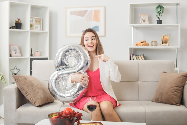 Verrast vrouw op happy women's day holding en wijst naar nummer acht ballon zittend op de bank in de woonkamer