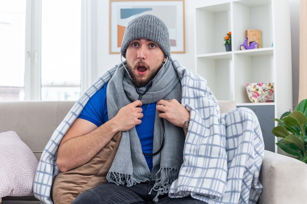 Verrast zieke jongeman met sjaal en muts met stethoscoop zittend op de bank in de woonkamer kijkend luisterend naar zijn eigen hartslag