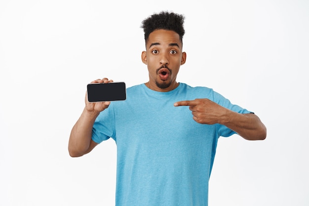 verraste afro-amerikaanse man hapt naar adem en zegt wauw, wijzende vinger naar het scherm van de mobiele telefoon met een verbaasd gezicht, met reclame op wit