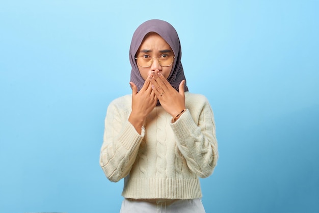 Verraste jonge aziatische vrouw die de mond bedekt met de hand voor een fout op een blauwe achtergrond