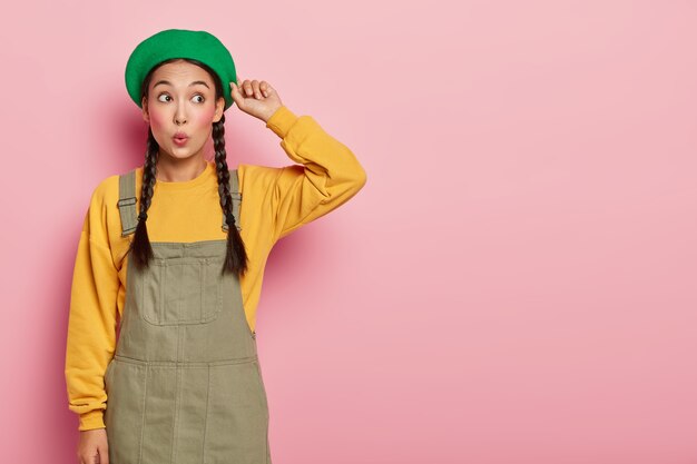 Verraste jonge Aziatische vrouw met afgeronde lippen, minimale make-up raakt baret en kijkt verbaasd opzij