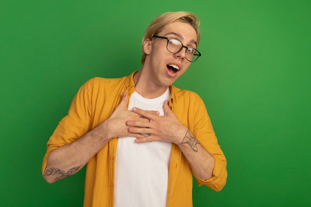 Verraste jonge blonde kerel die gele t-shirt en glazen draagt ?? die handen op hart zetten dat op groen met exemplaarruimte wordt geïsoleerd